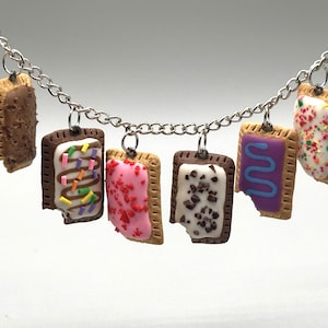 May include: A silver-toned metal chain necklace featuring six miniature rectangular pastry charms. Each charm resembles a toaster pastry with various icing colours, including brown, white with sprinkles, pink, white with dark bits, and purple.