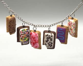 May include: A silver-toned metal chain necklace featuring six miniature rectangular pastry charms. Each charm resembles a toaster pastry with various icing colours, including brown, white with sprinkles, pink, white with dark bits, and purple.