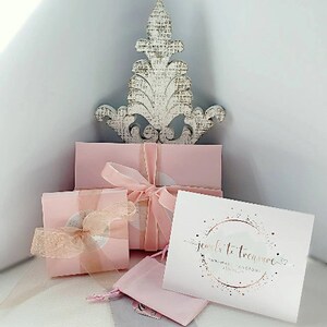 K&ouml;nnte beinhalten: Rosa Geschenkboxen mit einem wei&szlig;en Dekost&uuml;ck oben drauf. Die Boxen sind mit rosa Schleife gebunden und haben ein wei&szlig;es Etikett mit dem Text "jewels to treasure handmade custom jewelry" in goldenen Buchstaben.