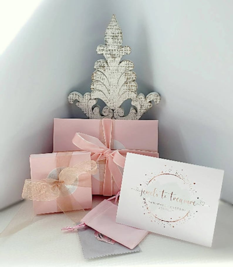 K&ouml;nnte beinhalten: Rosa Geschenkboxen mit einem wei&szlig;en Dekost&uuml;ck oben drauf. Die Boxen sind mit rosa Schleife gebunden und haben ein wei&szlig;es Etikett mit dem Text "jewels to treasure handmade custom jewelry" in goldenen Buchstaben.