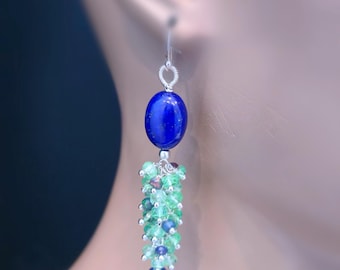 Könnte beinhalten: Ein silberner Ohrring mit einem großen, ovalen, tiefblauen Edelstein. Darunter hängt eine Ansammlung kleiner, grüner und blauer Perlen nach unten und erzeugt ein mehrschichtiges Design. Der Ohrring wird vor einem verschwommenen Hintergrund gezeigt.