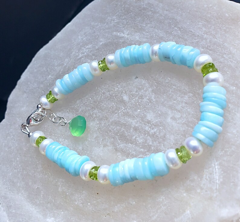 K&ouml;nnte beinhalten: Ein hellblaues und wei&szlig;es Perlenarmband mit gr&uuml;nen Edelstein-Akzenten. Das Armband hat einen silbernen Verschluss.