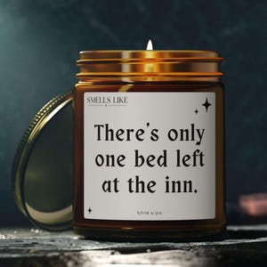 Op de afbeelding: Een brandende kaars in een amberkleurige glazen pot met een gouden deksel. Het witte etiket bevat de tekst "SMELLS LIKE" bovenaan, "There's only one bed left at the inn." in het midden en "BONNIE & MAC" onderaan, met kleine sterretjes.