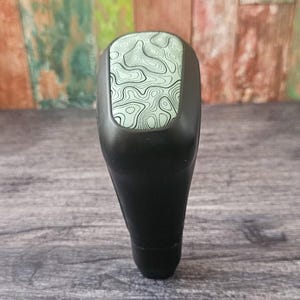 May include: A black gear shift knob with a light green and black topographic pattern on the top. The knob is smooth and rounded, with a sleek, modern design. The background is a wooden surface.