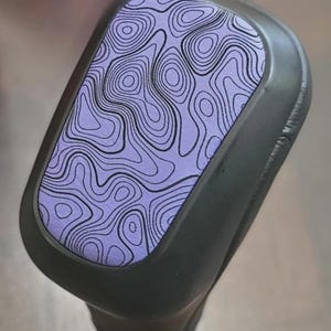 May include: A black object with a purple surface featuring a topographic map design. The map design is composed of black lines on a purple background. The object has a curved shape and appears to be made of a smooth material.