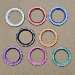 May include: Eight different colored metal rings with a white patterned design. The rings are arranged in a circle on a brown surface.