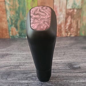 May include: Black car gear shift knob with a pink and black topographic pattern on the top. The gear shift knob is made of a smooth, solid material and is designed for easy installation.
