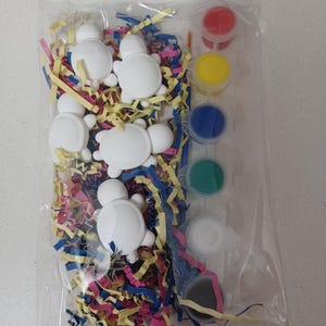 May include: A clear plastic bag containing five white turtle-shaped figurines nestled in colorful shredded paper filler, accompanied by a strip of six small paint pots in red, yellow, blue, green, white, and black, along with a thin paintbrush.
