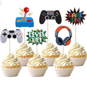 May include: Cupcakes with white frosting, decorated with gaming-themed toppers. Toppers include a joystick, game controllers, headphones, and text that says "Level Up!" and "Game On!" Perfect for a gamer's birthday party.