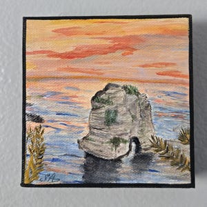 May include: Square canvas painting of a coastal scene at sunset. The artwork depicts a large rock formation in the sea, with a vibrant orange and pink sky. The water is blue, and the painting is framed in black.