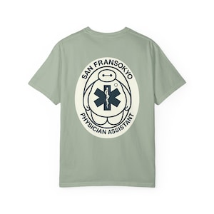 May include: Sage green t-shirt featuring a white oval graphic with the text "SAN FRANSOKYO PHYSICIAN ASSISTANT." The graphic includes a stylized medical symbol and a cartoon character design.