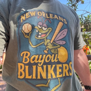 May include: Green t-shirt with a graphic featuring a cartoon insect holding a baseball bat and ball. The text on the shirt reads "New Orleans Bayou Blinkers" in yellow and blue.