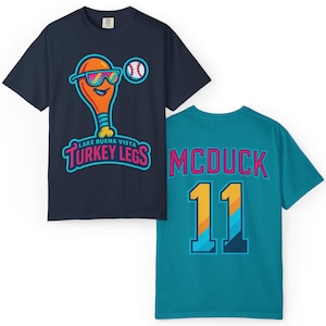 May include: Two t-shirts. One navy blue tee features a cartoon turkey leg wearing sunglasses and a baseball. The text reads "Lake Buena Vista Turkey Legs." The other is teal with "McDuck 11" on the back.