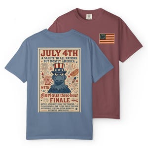 May include: Two short-sleeved t-shirts. One is dusty blue with a graphic on the back that reads "July 4th: A Salute to All Nations, But Mostly America." The other is maroon with a small flag graphic on the chest.
