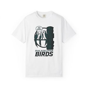 Puede incluir: Camiseta blanca con un diseño gráfico verde y negro. El diseño incluye tres loros, una máscara tribal y el texto "GO BIRDS!" y "PHILADELPHIA BIRDS". La camiseta tiene cuello redondo y mangas cortas.