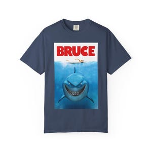 Op de afbeelding: Marineblauw T-shirt met een filmposterontwerp. Het woord "BRUCE" staat in rode blokletters boven een lachende blauwe haai in het water, met een zwemmer erboven. Een leuke, grafische tee.