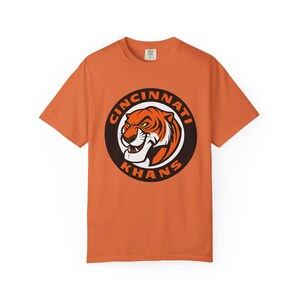 May include: Orange t-shirt featuring a circular graphic with a tiger head and the words "Cincinnati Khans" in a brown and white design. The shirt is a solid orange color.