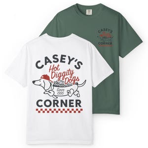 May include: Two t-shirts, one white and one green, featuring the text "Casey's Hot Diggity Dogs Corner" with a cartoon dachshund wearing a red hat. The white shirt has the full design on the back, while the green shirt has a smaller version on the upper back.