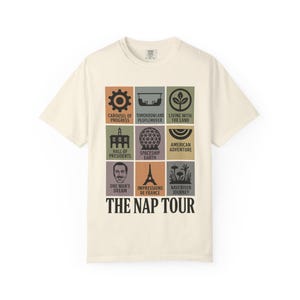 May include: Cream-colored t-shirt featuring a grid of colorful squares with themed icons and text. The shirt's design includes icons and text such as "Carousel of Progress," "Spaceship Earth," and "The Nap Tour."