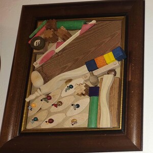 May include: A framed wooden art piece featuring a whimsical, colorful design. The artwork includes a pool ball, wooden shapes, and small figures, all set against a light background. The frame is a dark brown color.