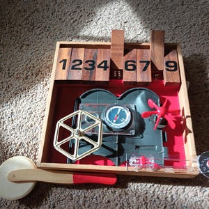 May include: A wooden toy with a film reel design. The toy features a black film reel, a red propeller, and a compass. The numbers 1, 2, 3, 4, 6, 7, and 9 are displayed on wooden blocks. A wooden handle with a red tip is also included.