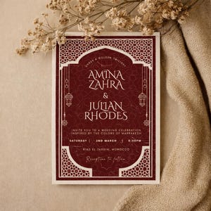 Puede incluir: Una invitación de boda burdeos con bordes y texto decorativos blancos. La invitación dice "Amina Zahra & Julian Rhodes" e invita a los invitados a una celebración de boda en Marruecos. Flores secas y tela beige en el fondo.