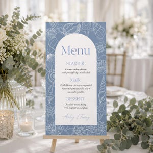 Pode incluir: Um cartão de menu retangular com um fundo azul poeirento apresentando ilustrações de linhas brancas de conchas e estrelas do mar. O centro tem um painel arqueado branco com o texto Menu, Starter, Main, Dessert e os nomes Audrey & Tommy.