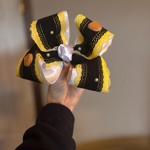 May include: A large, decorative hair bow featuring black fabric with yellow lace trim and white satin ribbon accents. The bow is adorned with small, round pearls and wooden embellishments. The bow is held in a hand.