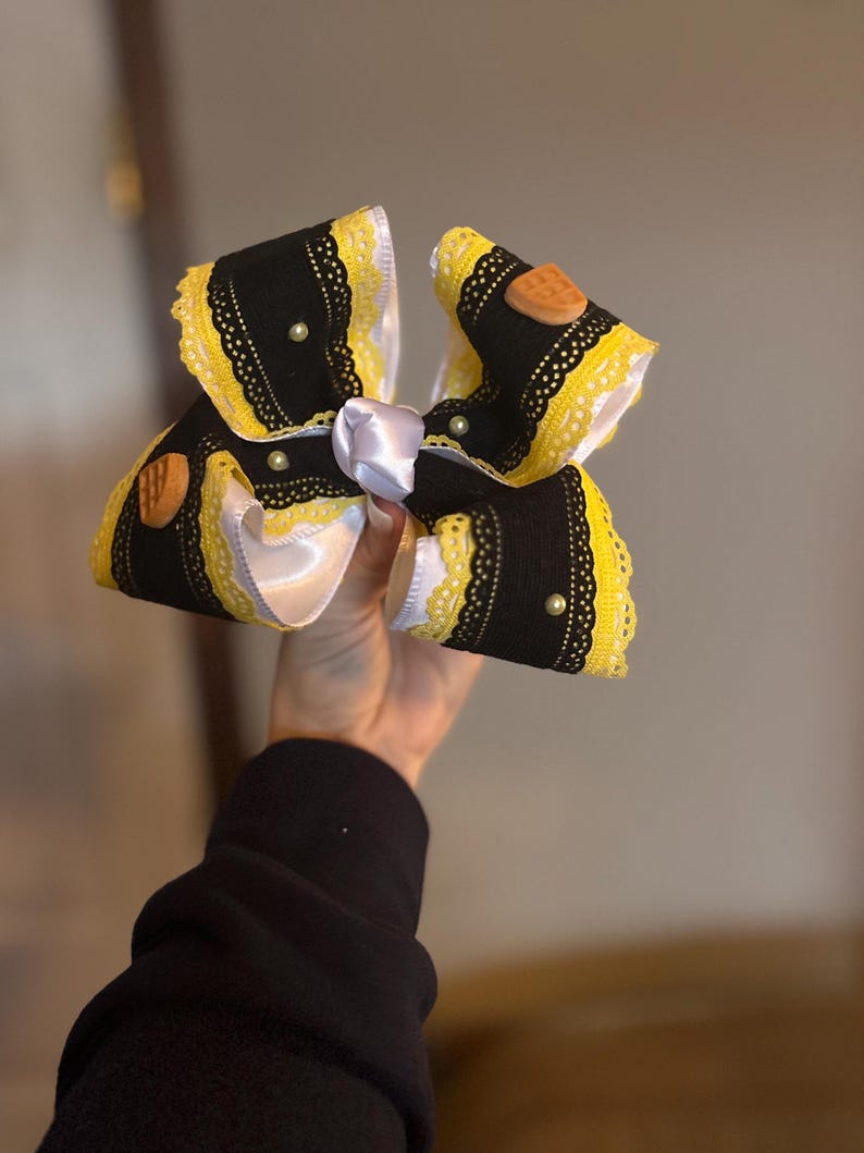 May include: A large, decorative hair bow featuring black fabric with yellow lace trim and white satin ribbon accents. The bow is adorned with small, round pearls and wooden embellishments. The bow is held in a hand.