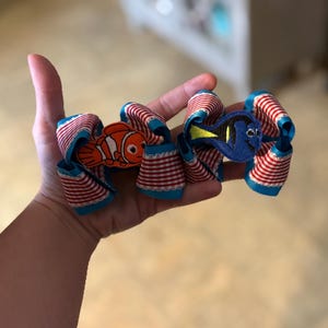 May include: A hair accessory featuring two loops of red and white striped ribbon with teal edges. An orange and white clownfish patch is attached to the left loop, and a blue and yellow tang fish patch is attached to the right loop of the bow.