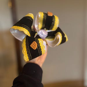 May include: A large, decorative hair bow featuring black and yellow ribbons with white lace trim. The bow has a waffle-shaped embellishment and a white satin center. The bow is held in a person's hand.