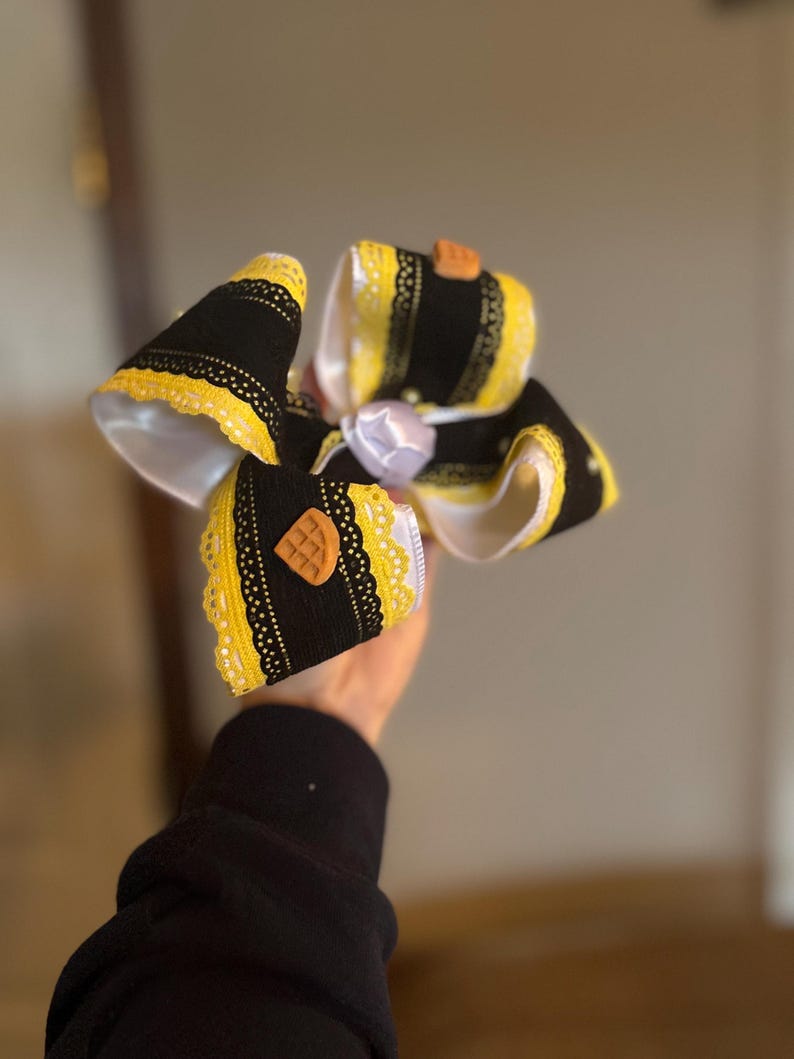 May include: A large, decorative hair bow featuring black and yellow ribbons with white lace trim. The bow has a waffle-shaped embellishment and a white satin center. The bow is held in a person's hand.