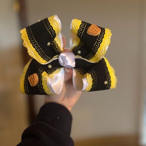 May include: A large, decorative hair bow featuring black fabric with yellow lace trim. The bow has a white satin center and is adorned with small pearls and waffle-shaped embellishments. The bow is held up against a neutral background.