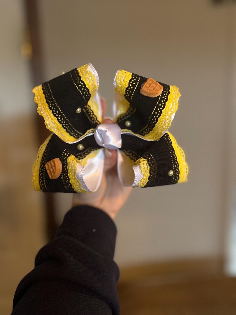 May include: A large, decorative hair bow featuring black fabric with yellow lace trim. The bow has a white satin center and is adorned with small pearls and waffle-shaped embellishments. The bow is held up against a neutral background.