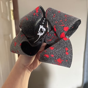 May include: A large hair bow featuring a dark gray glittery fabric base with a vibrant red paint splatter pattern. The bow is constructed with multiple loops and includes a black and white graphic detail of a knife on the central knot section.