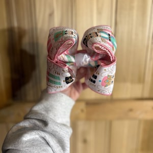 May include: A large hair bow made from white fabric patterned with pink, teal, and black designs. The bow has pink edging and a white centre knot. A decorative accessory to add a touch of flair to any hairstyle.