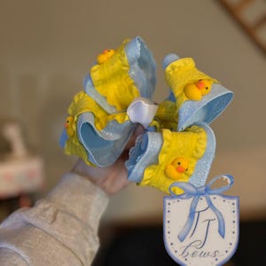 May include: A large, handmade hair bow featuring yellow and light blue fabric with a decorative duckling. The bow has a white center and is embellished with a blue ribbon. A tag with the text "TJ bows" is attached.