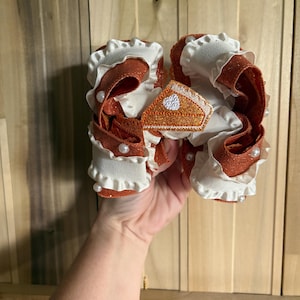 May include: A large hair bow featuring layers of orange glitter ribbon and white ruffled ribbon. The bow is accented with small white faux pearls and a central triangular patch shaped like a slice of pumpkin pie with a white heart detail.