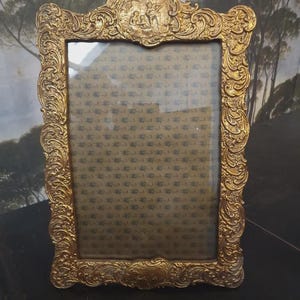 Puede incluir: Marco de fotos dorado ornamentado con intrincados detalles de volutas y una abertura rectangular. Una pequeña escena en relieve adorna la parte superior. El marco está vacío, revelando un fondo estampado a través del cristal.