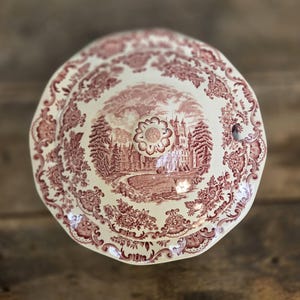 May include: A round ceramic tureen lid featuring a cream-coloured base with a detailed maroon transferware pattern. The design depicts a large castle surrounded by trees and a landscape, framed by an ornate floral and scrollwork border.