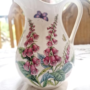 Puede incluir: Jarra de cerámica blanca con asa, decorada con flores de dedalera rosa, hojas verdes y mariposas. La jarra tiene un pico curvo y una abertura ancha, adecuada para servir bebidas.