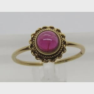 May include: A gold ring with a bright pink gemstone, set in a decorative, rope-style border. The ring's band is plain gold. The gemstone is approximately 1.3 cm in diameter, offering a classic and elegant design. The ring is a piece of jewellery.