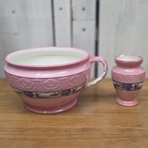 May include: A pink ceramic bowl with a handle and a matching small pink vase. Both pieces feature a white interior, a raised relief pattern around the upper rim, and a horizontal band decorated with a colorful landscape scene near the base.