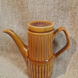 May include: A brown ceramic coffee pot with a lid. The pot has a long spout and a handle. The body of the pot is decorated with vertical lines. The lid has a floral design. The pot is made of ceramic.