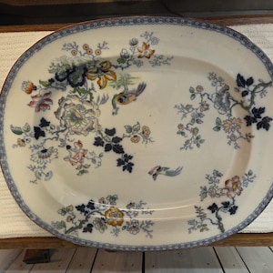 May include: An oval ceramic serving platter featuring a white base with a blue decorative border. The centre is adorned with a multi-coloured floral pattern, including dark blue leaves, orange blossoms, and two small birds in flight.