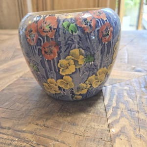 May include: A ceramic vase with a rounded shape, featuring a blue background adorned with orange and yellow floral patterns. The vase has a glossy finish and is likely used for decorative purposes.