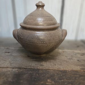 May include: A round, light brown ceramic lidded bowl with two side handles and a matching conical lid featuring a small knob handle. The surface has a speckled, textured finish with subtle variations in tone across the entire vessel.