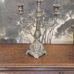 Peut inclure: Un candélabre en métal argenté doté de trois bougeoirs, d'une tige centrale ornée et d'une large base décorative. La pièce présente des volutes complexes et des motifs floraux en relief sur toute sa structure, avec une finition patinée.