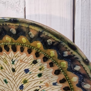 May include: Close-up of a decorative ceramic plate with a detailed, hand-painted design. The plate features a cream-coloured centre with a feather-like pattern, bordered by a band of brown, blue, and green. The rim has a scalloped edge.