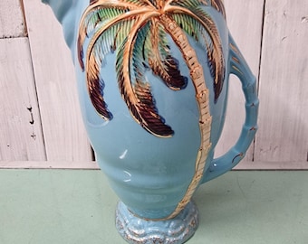 May include: A light blue ceramic pitcher with a palm tree design. The palm tree is detailed with brown, gold, and green accents. The handle has a bamboo-like design, and the base has a decorative, textured pattern.